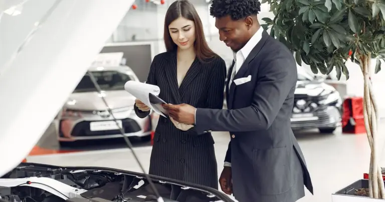 Vendedor e compradora conferem um documento juntos ao lado de um carro com o capô aberto
