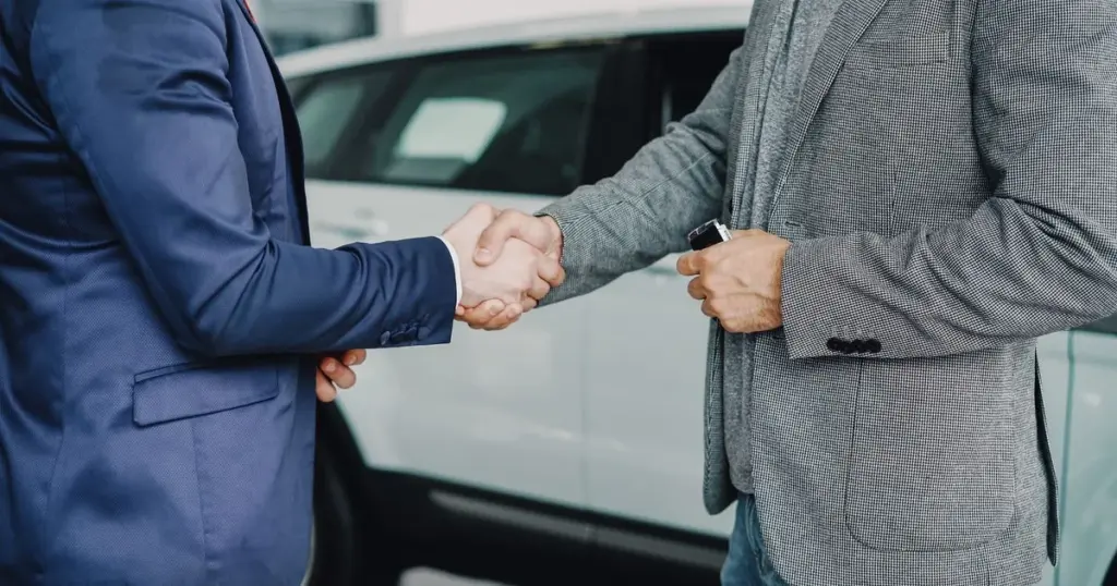 Comprador e vendedor apertam as mãos ao lado de um carro usado, fechando a negociação