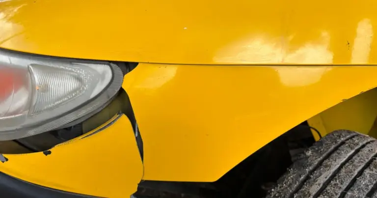 Detalhe de um para-choque amarelo amassado com farol danificado, mostrando avaria de colisão