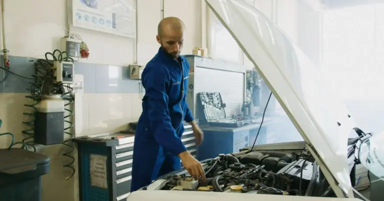 Mecânico de macacão azul inspeciona o motor de um carro com o capô aberto em uma oficina