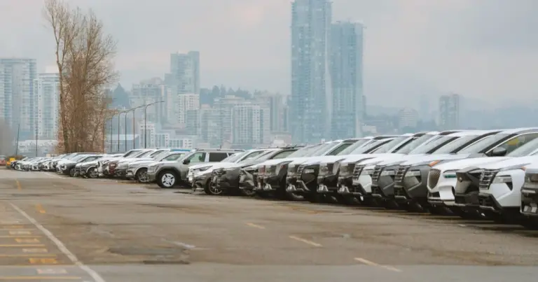 Vista de uma fileira longa de carros seminovos estacionados em um pátio, com prédios ao fundo