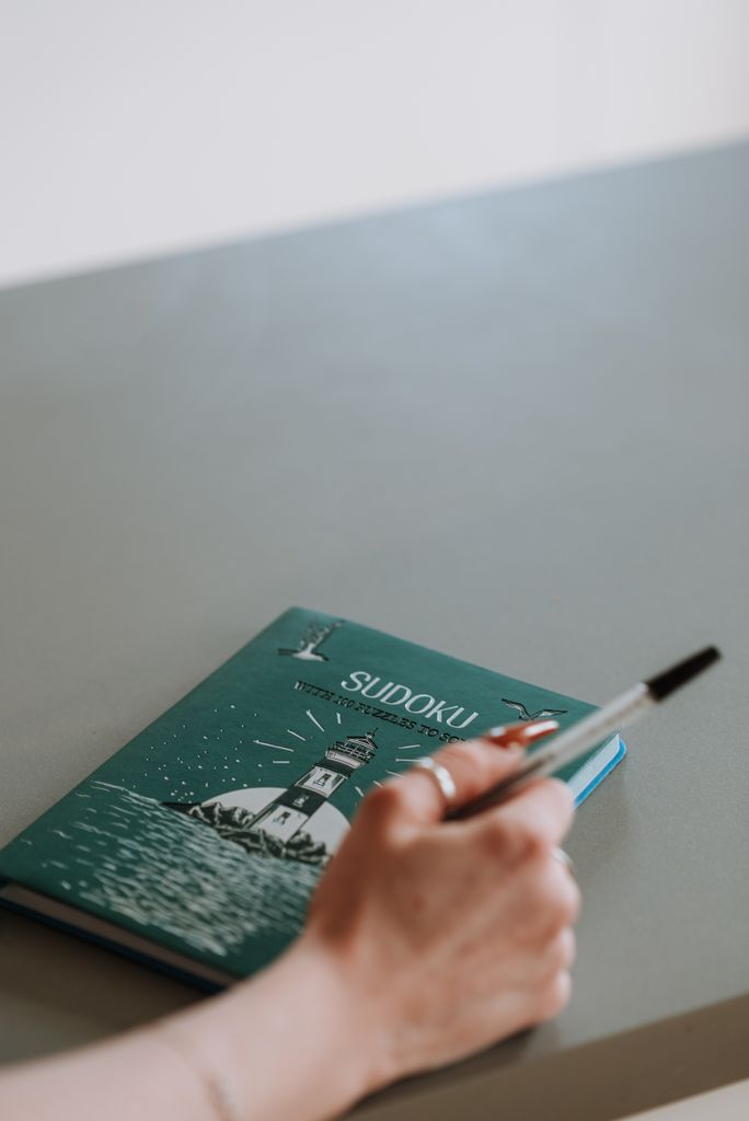 Mão segurando uma caneta ao lado de um livro de Sudoku sobre a mesa.