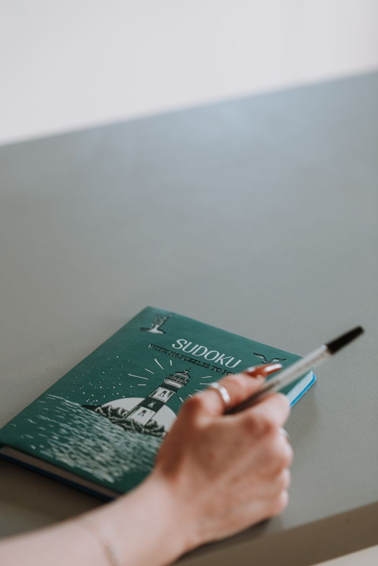 Mão segurando uma caneta ao lado de um livro de Sudoku sobre a mesa.